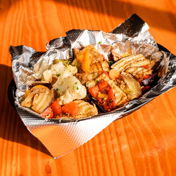 Grilled vegetables, including eggplant, cauliflower, and bell peppers, in a foil-lined container on a wooden table.
