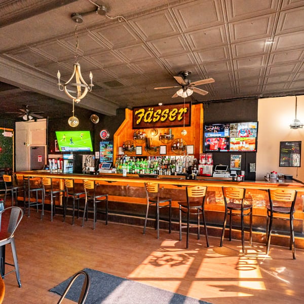 Interior of Fässer bar-&-grill with a long wooden bar, stools, and multiple TVs showing sports.