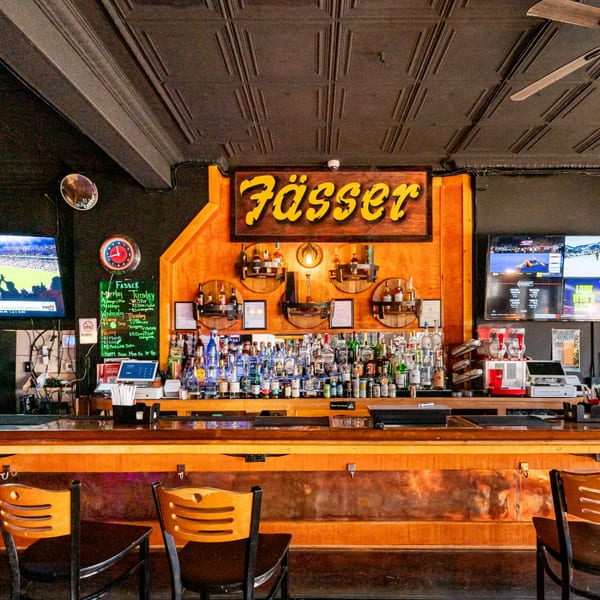 The Fässer bar interior, featuring a well-stocked liquor display, bar stools, and menu boards.