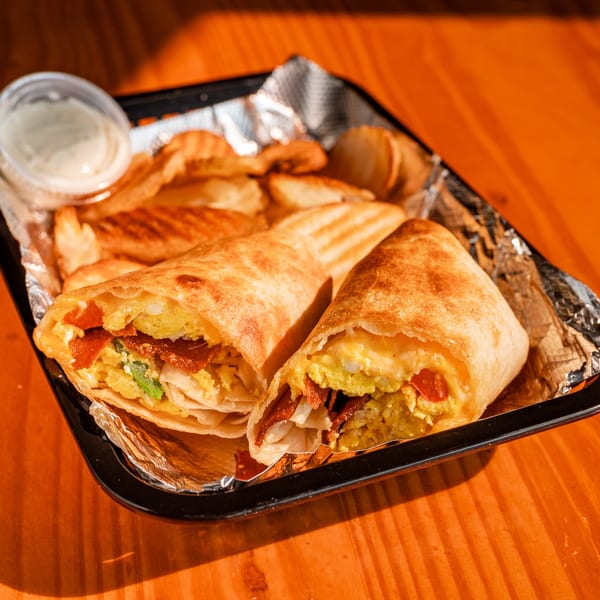 Two breakfast burritos with bacon, egg, and vegetables, served with potato wedges and dipping sauce.