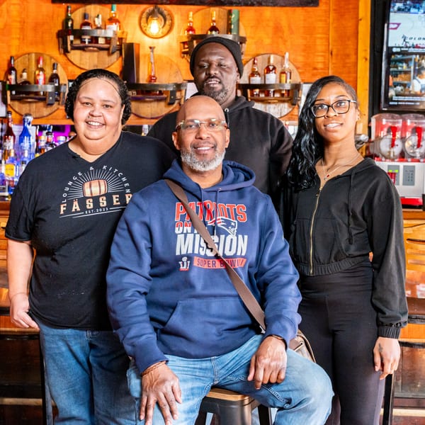 Four Fässer bar-&-grill staff members smiling in front of a wooden bar with liquor bottles.